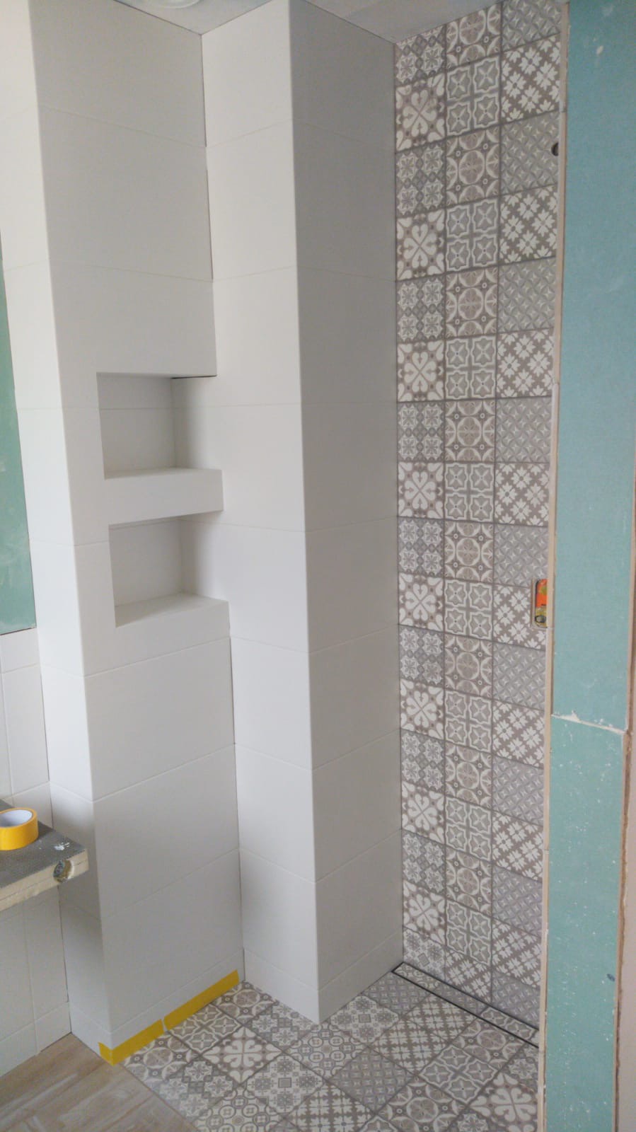 Wnętrze łazienki w trakcie remontu: białe płytki ścienne z wnękami, dekoracyjne szaro-białe płytki podłogowe i ścienne, widoczne taśmy malarskie i surowa płyta gipsowo-kartonowa.