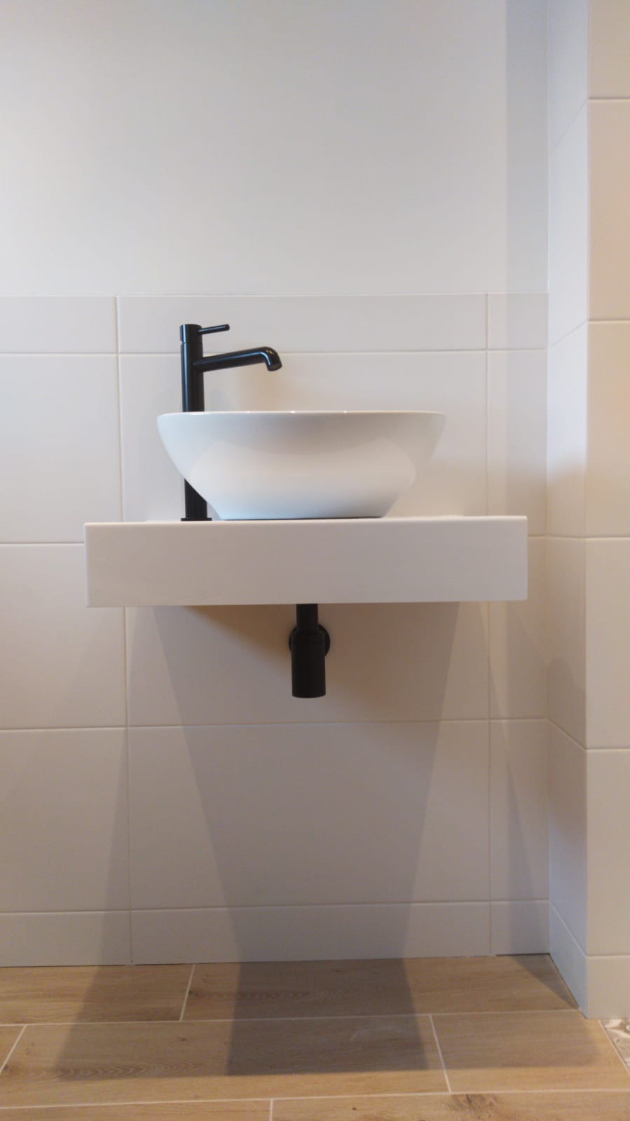 Nowoczesna, minimalistyczna łazienka z białą, okrągłą umywalką nablatową i czarną armaturą, zamontowana na białym blacie. Ściany wyłożone jasnymi, kwadratowymi płytkami, a podłoga panelami...