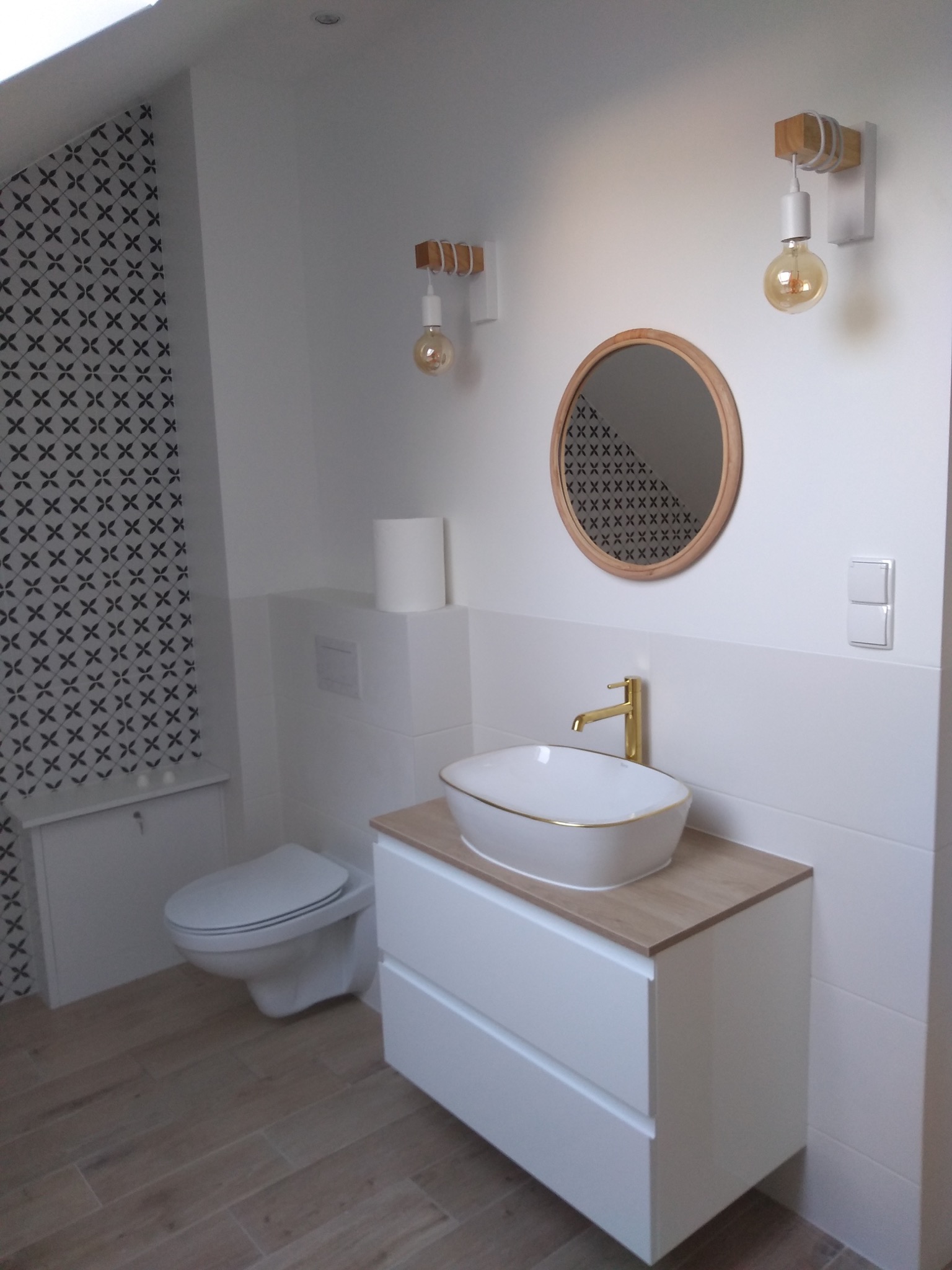 Nowoczesna łazienka z białą umywalką nablatową z delikatnym złotym rantem, złotą baterią, okrągłym lustrem w drewnianej ramie i dekoracyjną tapetą w geometryczny wzór, oświetlona dwoma lampami...