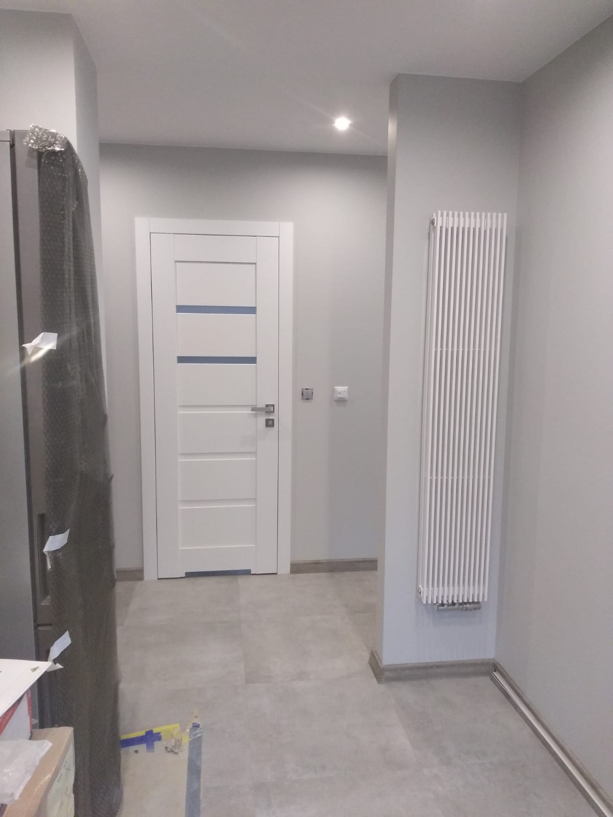 Wykończone wnętrze przedpokoju w odcieniach szarości z białą stolarką drzwiową z poziomymi, niebieskimi akcentami i grzejnikiem panelowym.