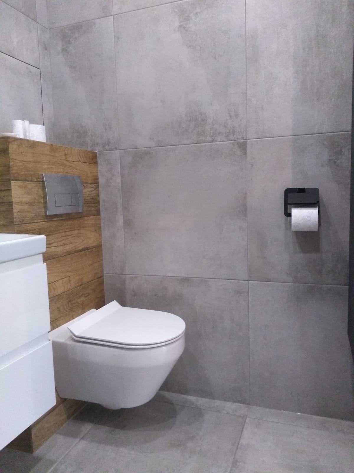 Nowoczesna łazienka z szarymi płytkami imitującymi beton, podwieszana toaleta z deską slim, drewnopodobna zabudowa stelaża i biała szafka pod umywalką.