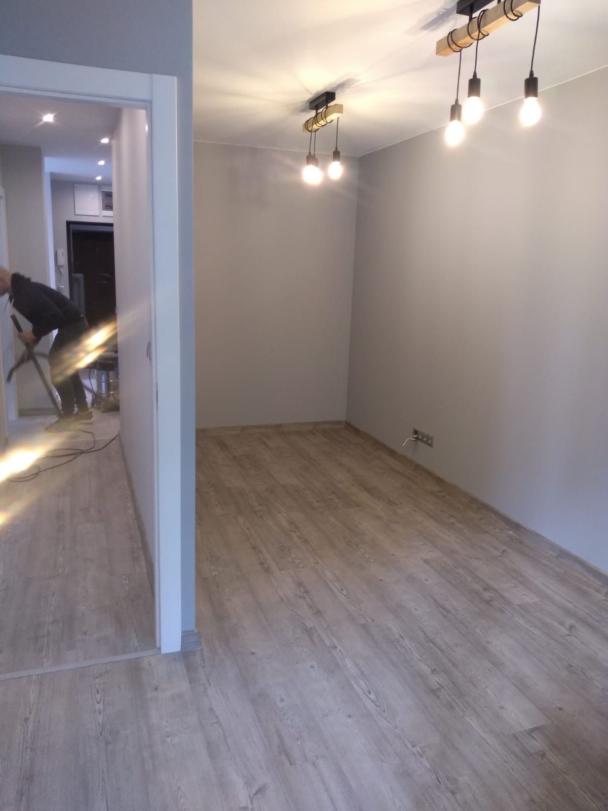 Pokój po remoncie z nową podłogą z paneli, widoczne lampy sufitowe z żarówkami i osoba odkurzająca w tle.