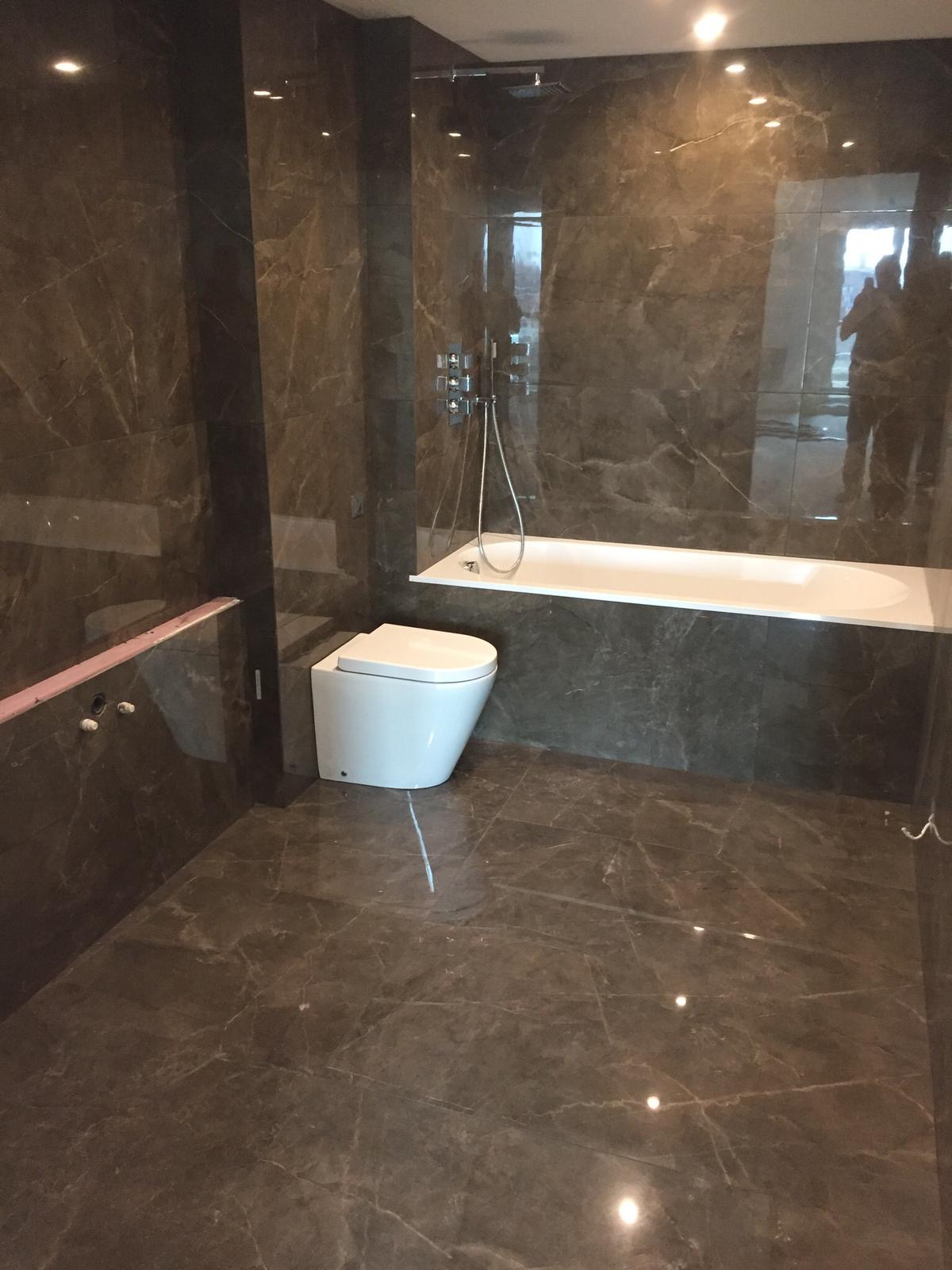 Nowoczesna łazienka z ciemnymi, marmurkowymi płytkami na ścianach i podłodze, białą wanną i podwieszaną toaletą; widoczne chromowane elementy armatury.