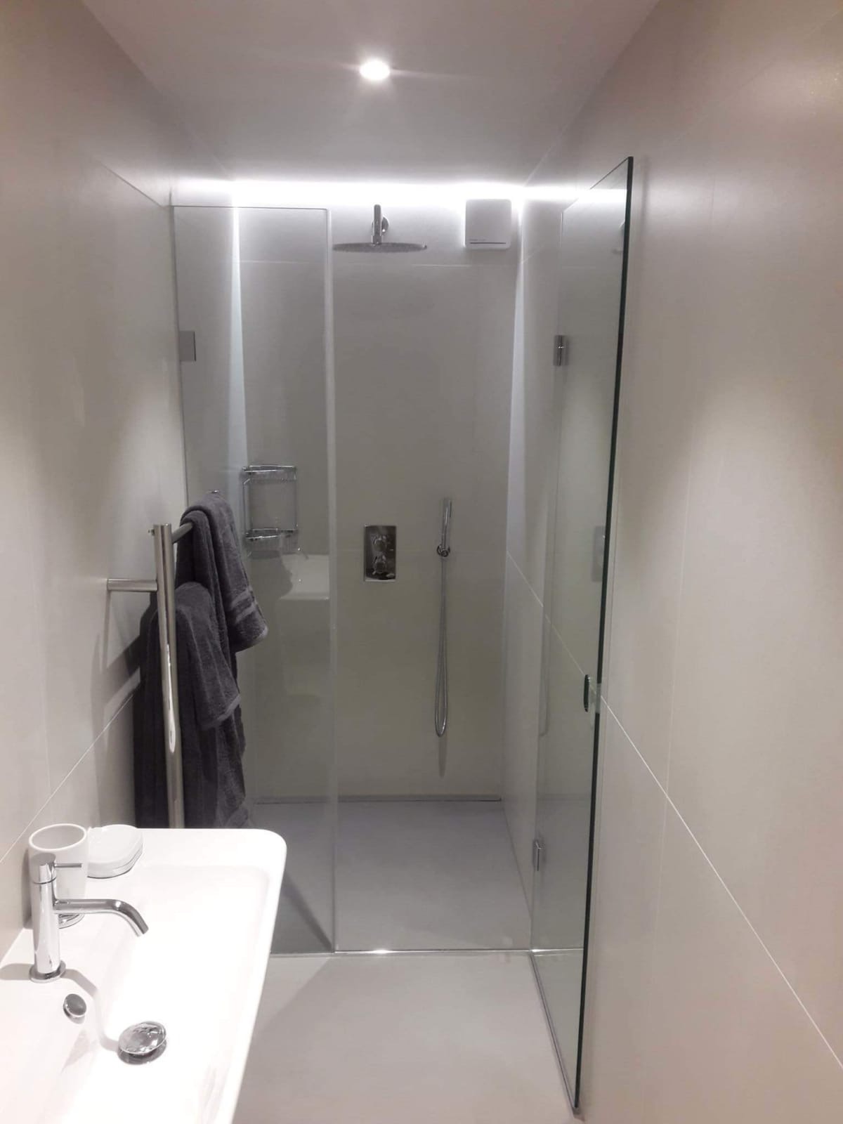 Nowoczesna łazienka z przeszkloną kabiną prysznicową typu walk-in, chromowaną armaturą i grzejnikiem drabinkowym z szarymi ręcznikami, oświetlona punktowym światłem sufitowym i paskiem LED nad kabiną.