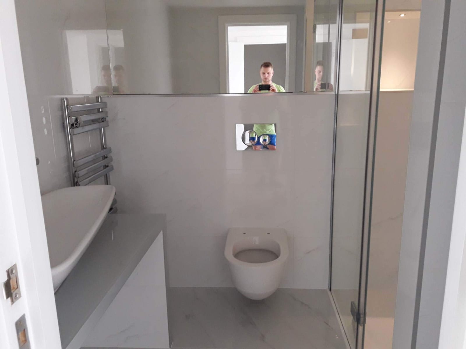 Nowoczesna łazienka z białymi płytkami imitującymi marmur, podwieszana toaleta, grzejnik drabinkowy, umywalka nablatowa na szarym blacie i kabina prysznicowa ze szklanymi drzwiami; widoczne odbicie...