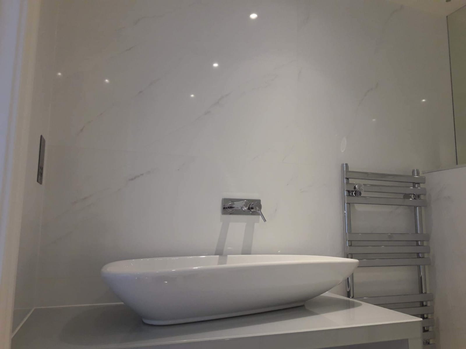 Biała, owalna umywalka nablatowa z chromowaną baterią na tle ściany wyłożonej białymi płytami imitującymi marmur, widoczny grzejnik drabinkowy i punktowe oświetlenie.