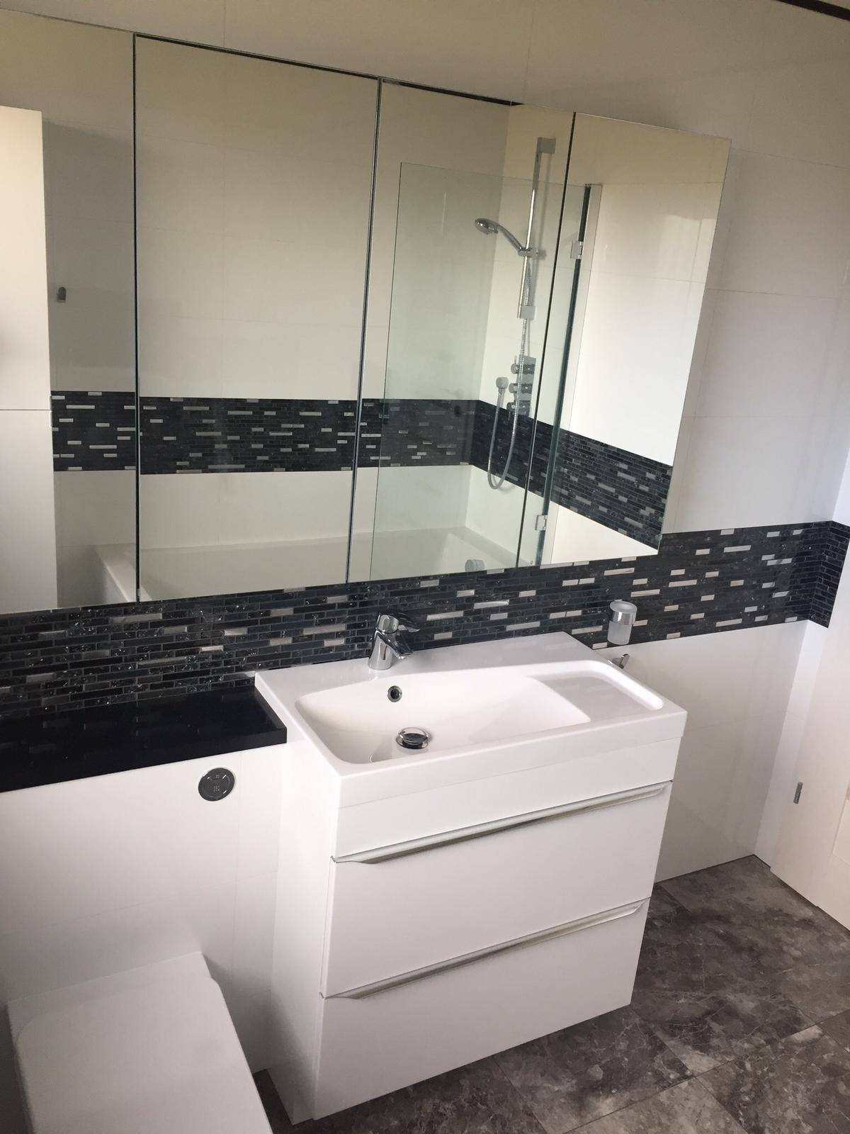Nowoczesna łazienka z białą umywalką z szafką, czarnym blatem i lustrzaną szafką wiszącą, ozdobiona pasem czarno-białej mozaiki, z widocznym fragmentem kabiny prysznicowej w odbiciu.
