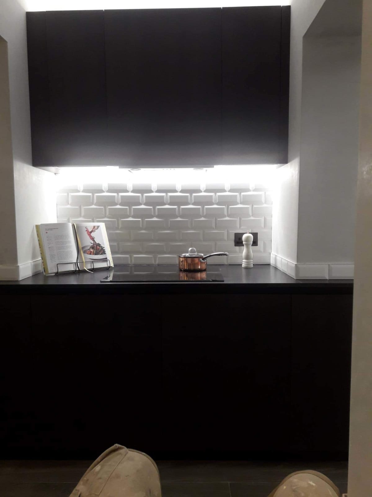 Nowoczesna kuchnia z czarnymi szafkami, białymi płytkami typu metro, oświetleniem LED pod szafkami, otwartą książką kucharską i miedzianym rondlem na płycie indukcyjnej.