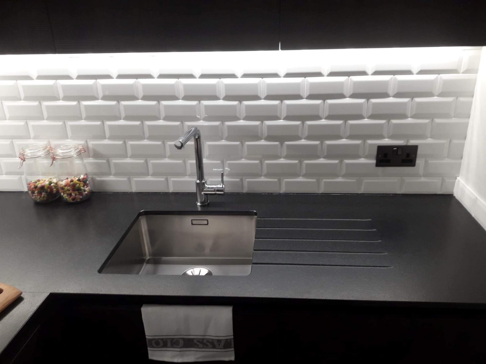 Nowoczesna kuchnia z czarnym blatem, stalowym zlewem, baterią i białymi kafelkami typu metro. Na blacie stoją dwa słoiki z kolorowymi cukierkami. Na szafce pod zlewem wisi biała ściereczka.