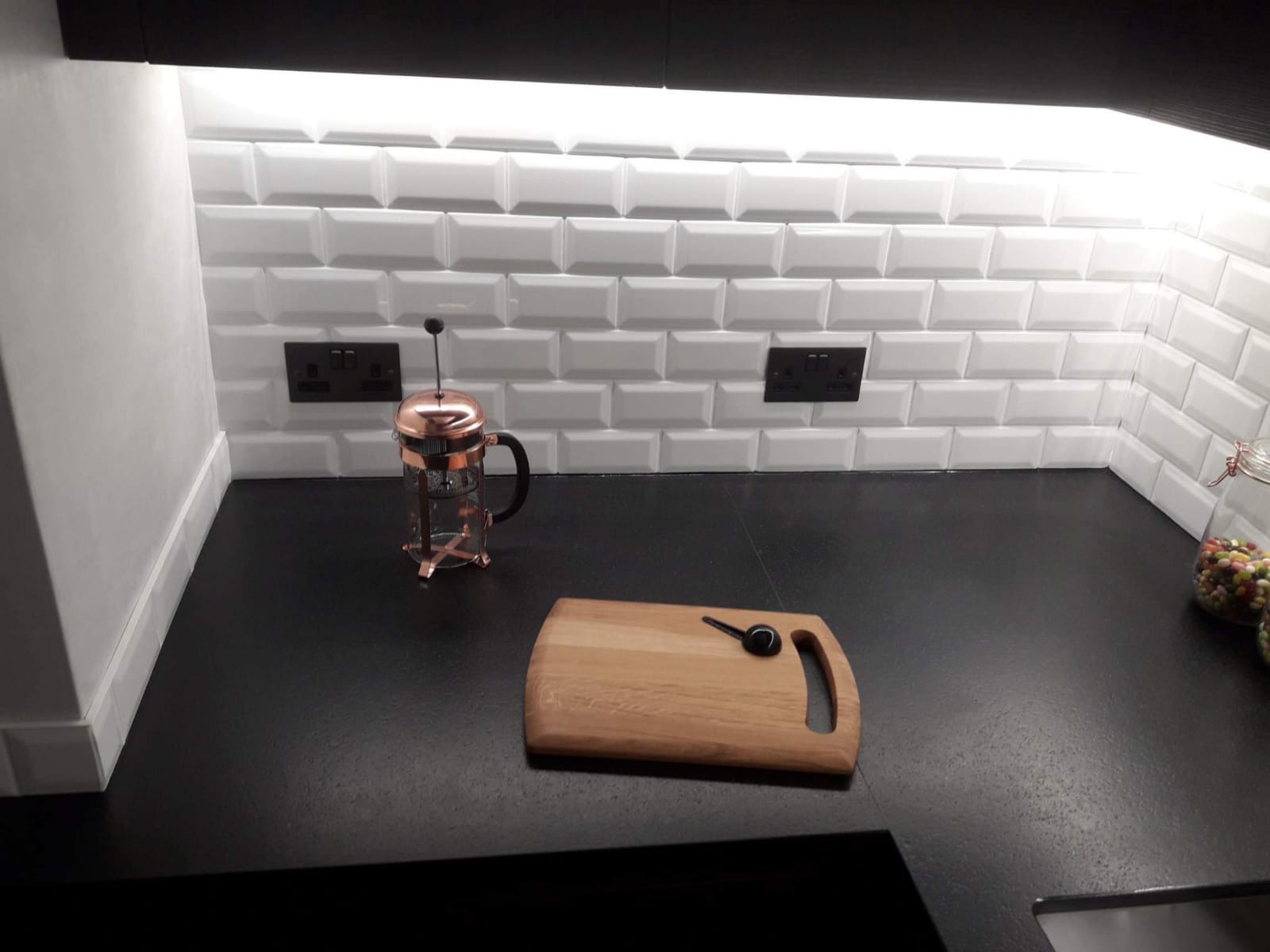Białe, kafelkowe płytki metra z czarną fugą i czarnymi gniazdkami elektrycznymi nad czarnym blatem kuchennym, na którym leży drewniana deska do krojenia z czarną łyżeczką i miedziany zaparzacz...