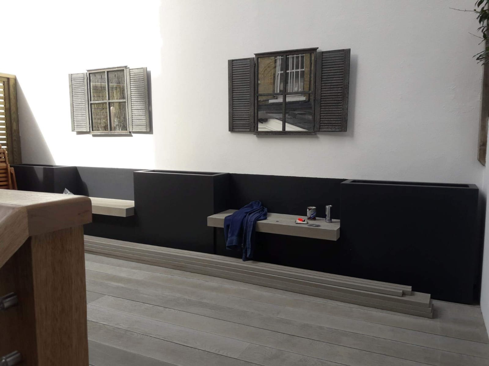 Wnętrze balkonu w trakcie wykańczania: widoczne czarne donice, drewniane panele podłogowe, dekoracyjne okna na ścianie, deski podłogowe do ułożenia oraz narzędzia malarskie na półce.