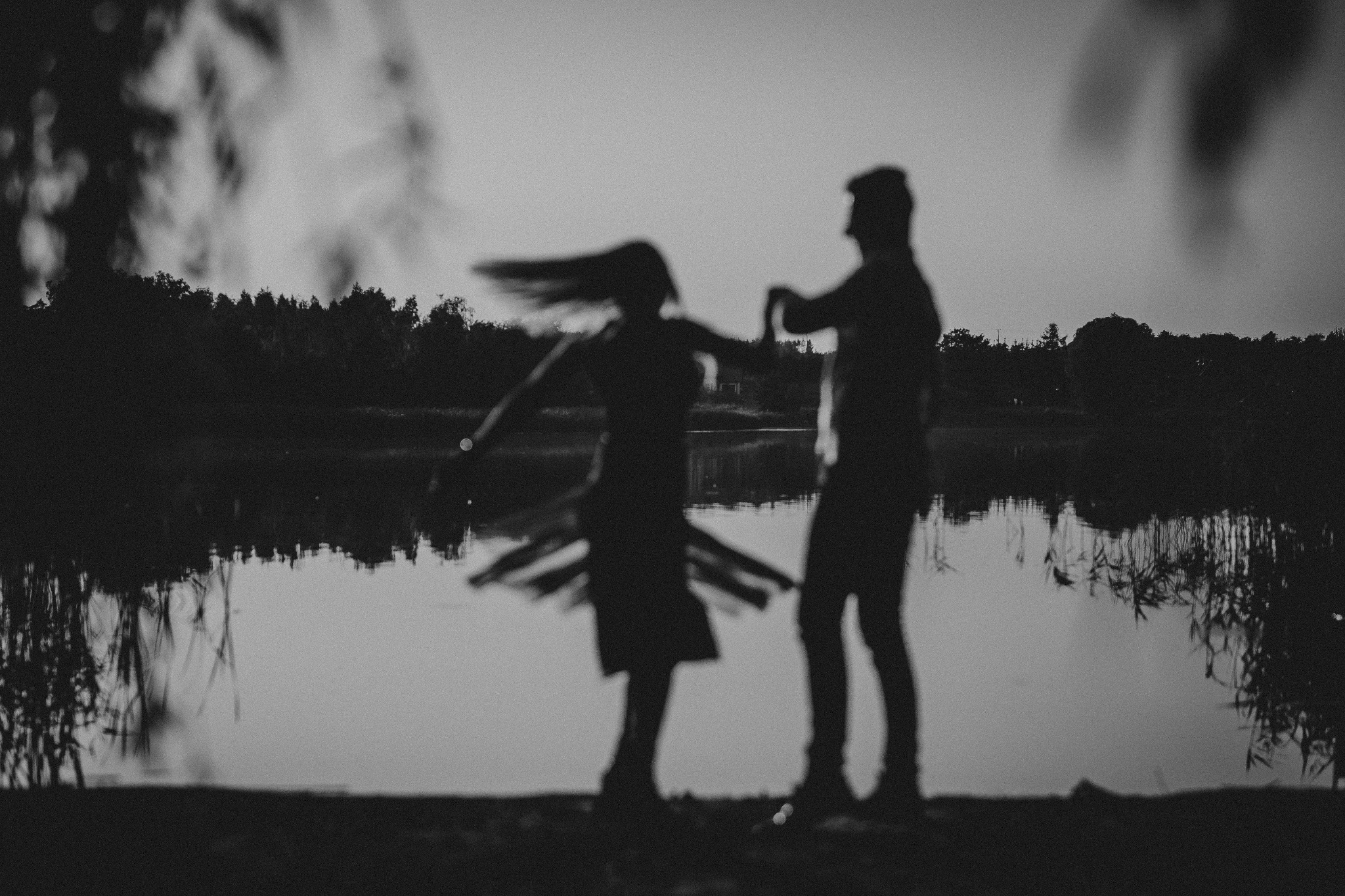Czarno-biała fotografia sylwetek pary tańczącej nad brzegiem jeziora, z rozmytym tłem drzew i odbiciem w wodzie.