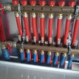 Rozdzielacz podłogowy z ośmioma obwodami, rury czerwone i niebieskie, zawory odcinające na wejściu, numery obwodów nadrukowane na belce.