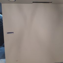 Mariusz Bartocha - Świeżo pomalowana szara ściana z widocznymi śladami taśmy malarskiej, zakrytymi gniazdkami i narzędziami malarskimi na podłodze.