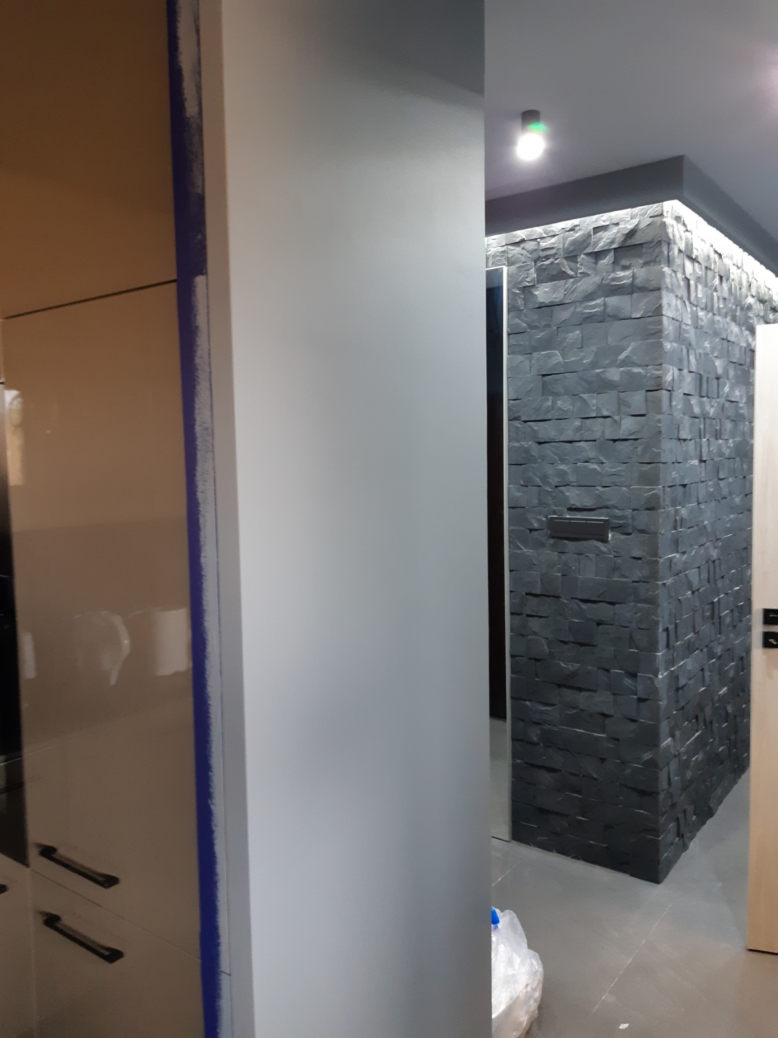 Fragment korytarza z dekoracyjną ścianą z ciemnego kamienia podświetloną ledami, widoczny fragment szafy z niebieskim zabezpieczeniem krawędzi i biała ściana.