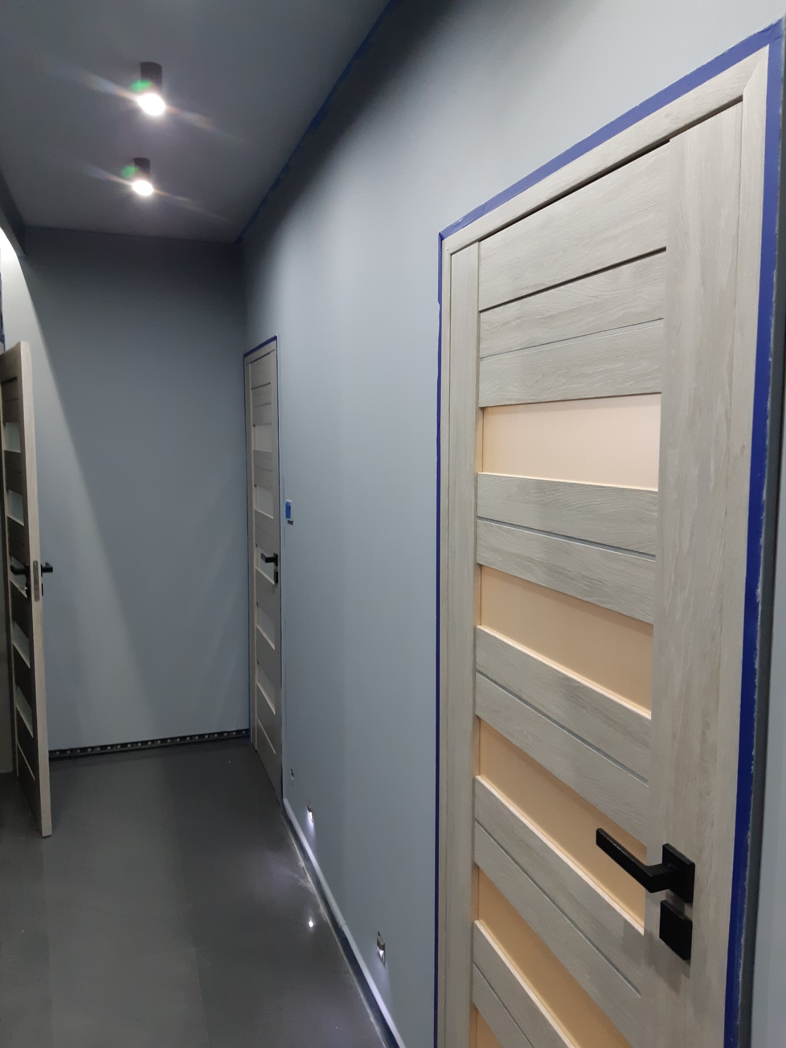 Korytarz w odcieniach szarości z trzema drewnianymi drzwiami z poziomymi panelami i czarnymi klamkami, widoczne taśmy malarskie przy futrynach i suficie, punktowe oświetlenie podłogowe i sufitowe.