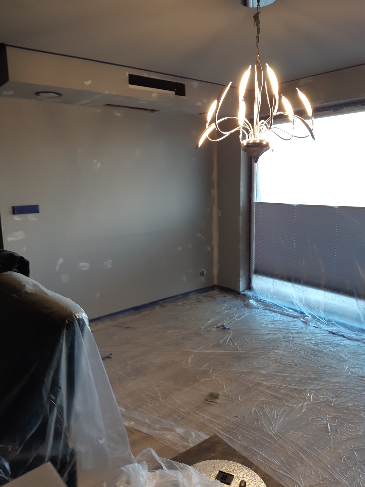 Przygotowanie do malowania w Zgierzu: pokój zabezpieczony folią malarską, ściany z widocznymi punktami po szpachlowaniu, nowoczesny żyrandol w centrum kadru.