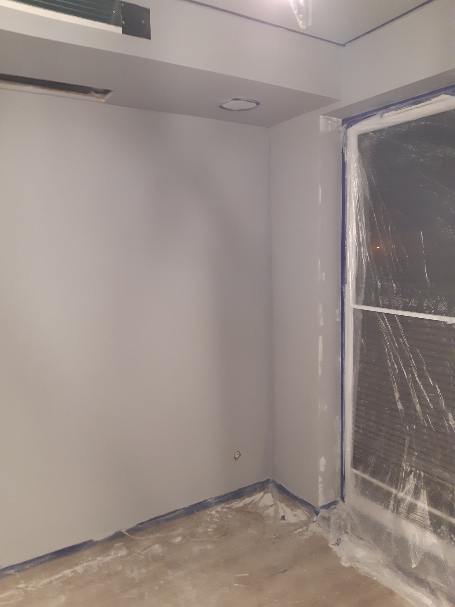 Pokój w trakcie odświeżania: ściany pomalowane na szaro, okno zabezpieczone folią malarską, widoczne ślady farby na podłodze i taśma malarska przy podłodze.