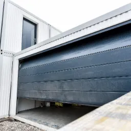 Szary segmentowy garaż wbudowany w białą konstrukcję modułową, częściowo otwarty, widoczne wnętrze garażu z narzędziami.