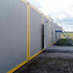 Zewnętrzny widok długiego, szarego budynku z modułów z żółtymi akcentami, z wejściem i schodami na kółkach, na tle zieleni i budynków w oddali.