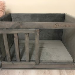 Klaudia Mościńska - Szare, tapicerowane legowisko dla psa z drewnianym frontem, ozdobione wygrawerowanym logo 'GUTEK' i symbolem domu z sercem, na jasnej podłodze.