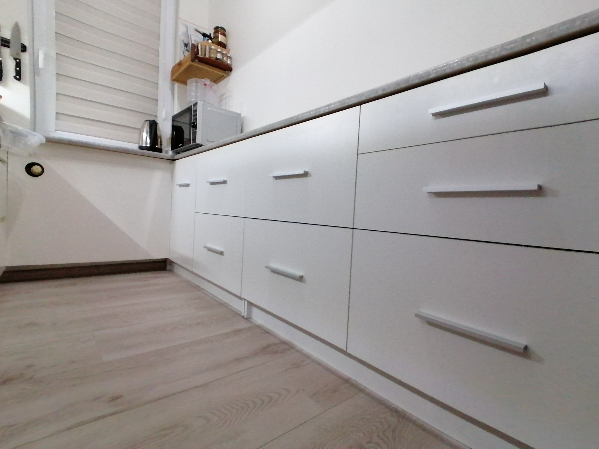 Biała zabudowa kuchenna z szufladami i szafkami, widok z niskiego kąta, prezentująca minimalistyczny design z podłużnymi uchwytami i blatem w odcieniach szarości.