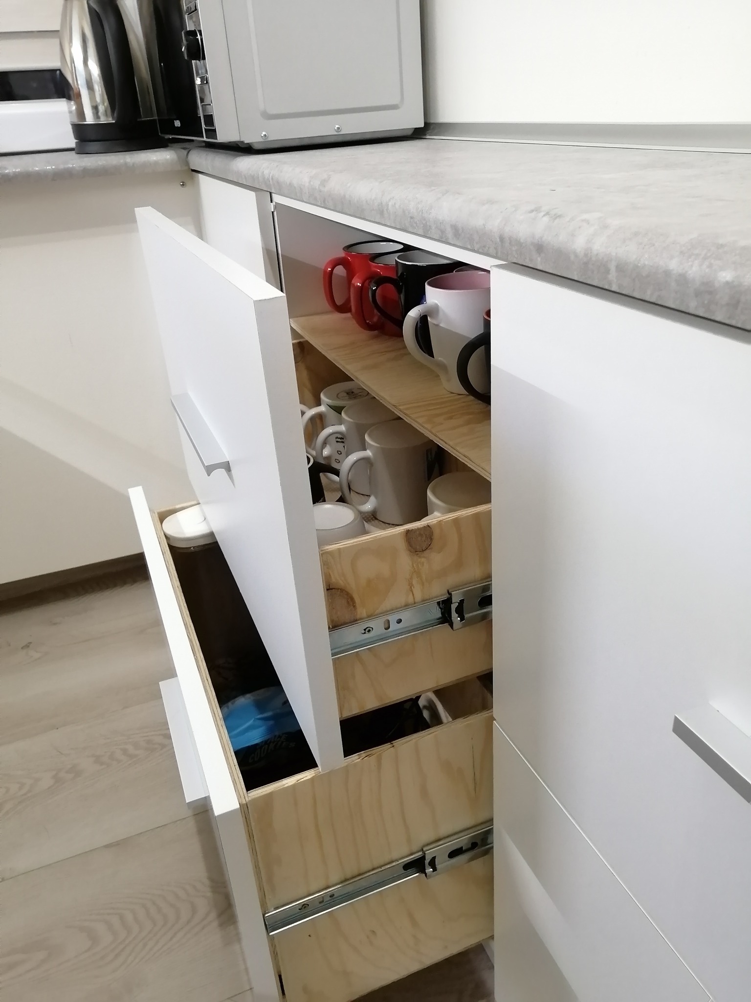 Wysuwane szuflady kuchenne z drewnianymi wkładami, z różnymi kubkami i pojemnikami, zamontowane w białej zabudowie kuchennej pod jasnym blatem, z widocznymi prowadnicami szuflad.