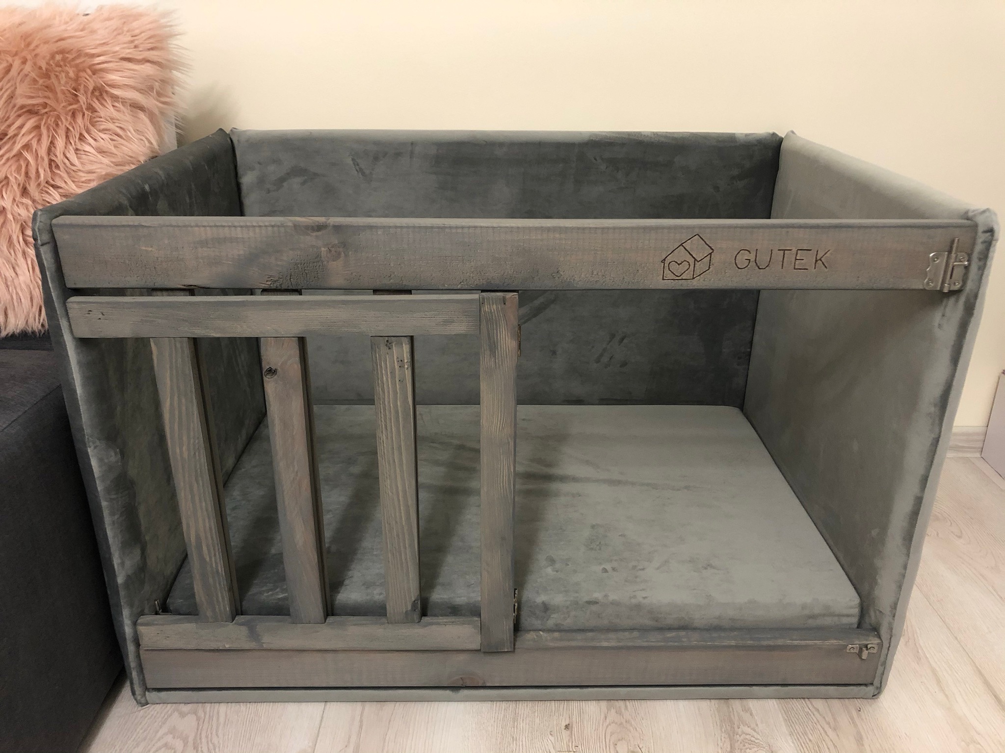 Szare, tapicerowane legowisko dla psa z drewnianym frontem, ozdobione wygrawerowanym logo 'GUTEK' i symbolem domu z sercem, na jasnej podłodze.