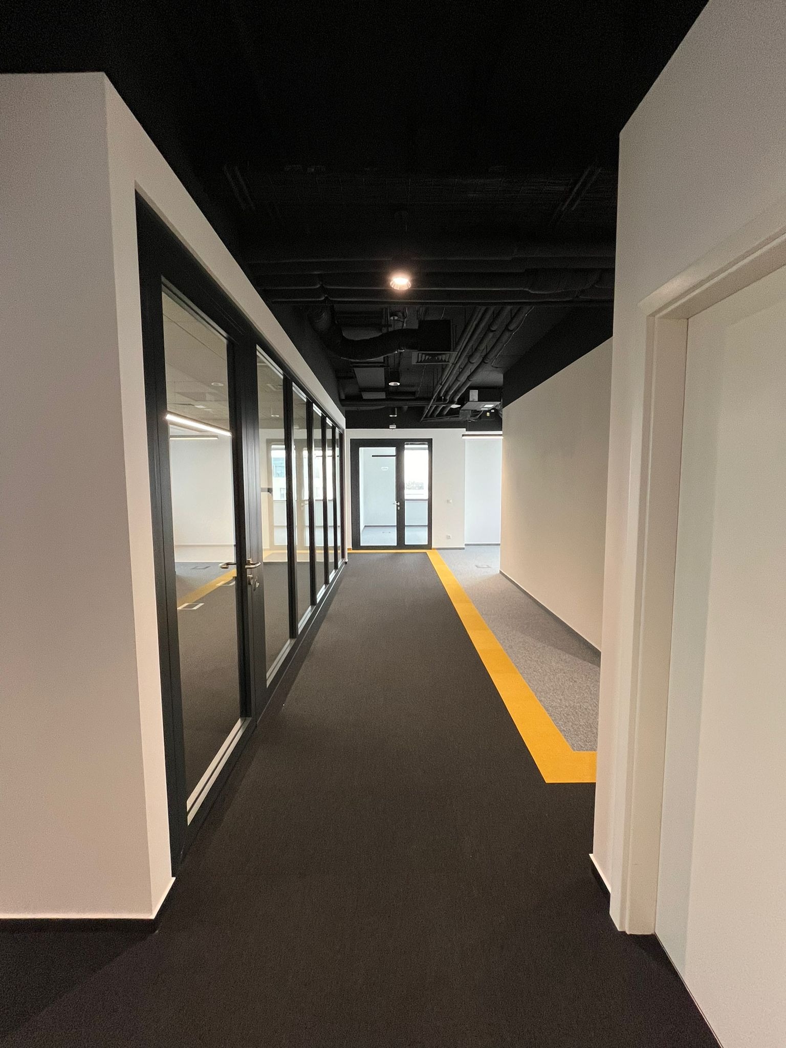Korytarz biurowy z czarną wykładziną i żółtą linią bezpieczeństwa, przeszklone biura po lewej stronie, sufit w kolorze czarnym z widocznymi instalacjami.