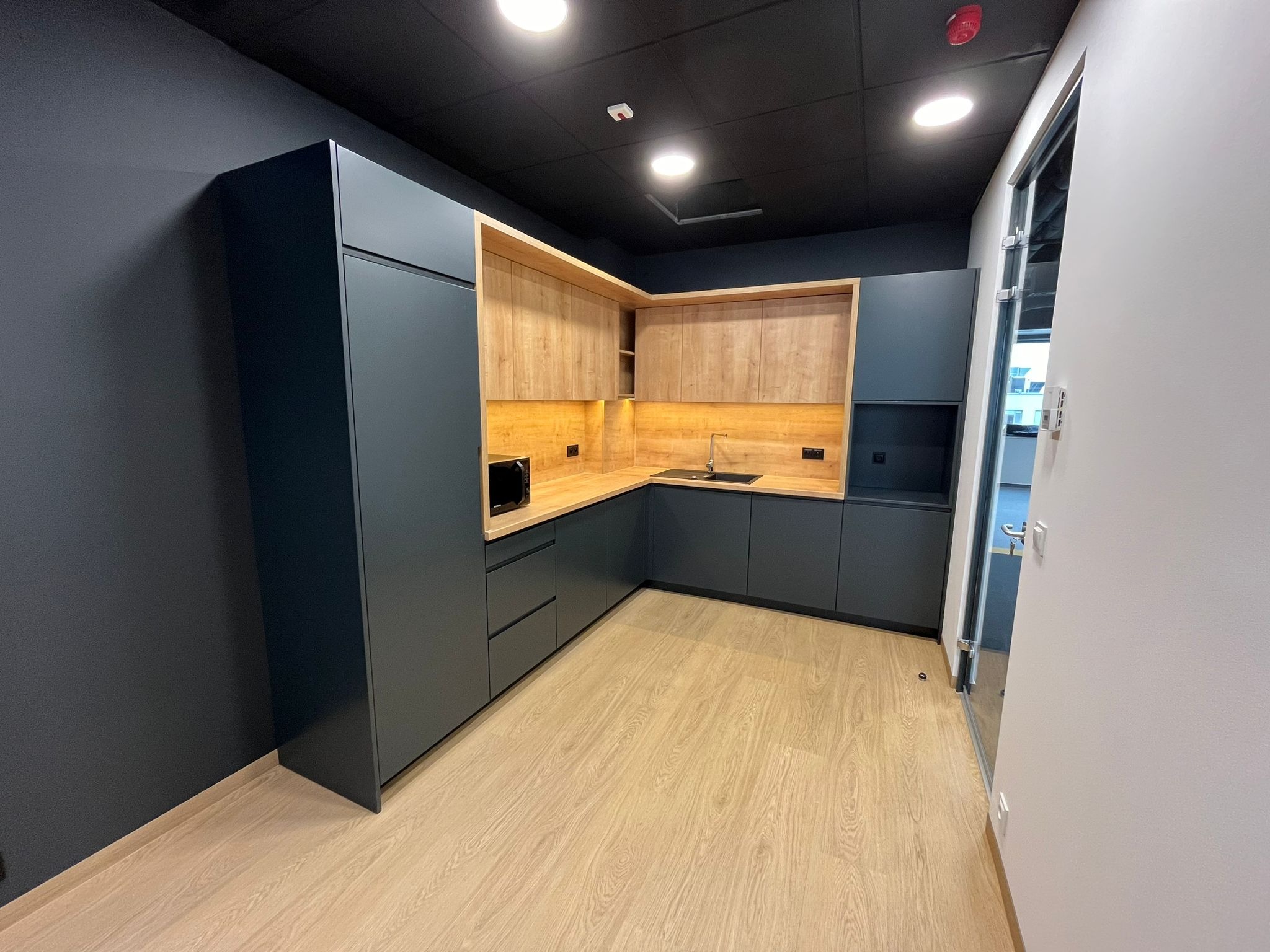 Nowoczesna kuchnia biurowa z ciemnoszarymi szafkami i drewnianymi blatami, oświetlona od spodu, z widocznym mikrofalówką i czarnym sufitem z punktowym oświetleniem.