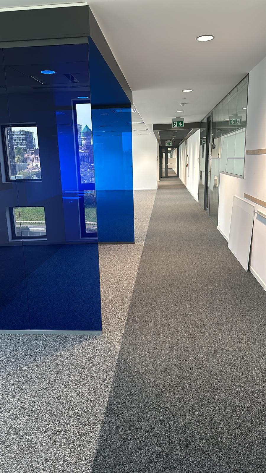 Długi korytarz biurowy z dwoma rodzajami wykładzin: szarą dywanową i jasnoszarą w plamki, oddzielonymi niebieską szklaną ścianką działową.
