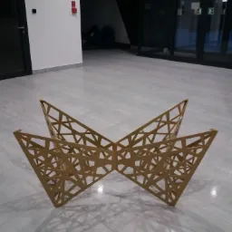 Geometryczna, ażurowa konstrukcja dekoracyjna z metalu w kształcie rozłożystego motyla, ustawiona na szarej podłodze w nowoczesnym wnętrzu.