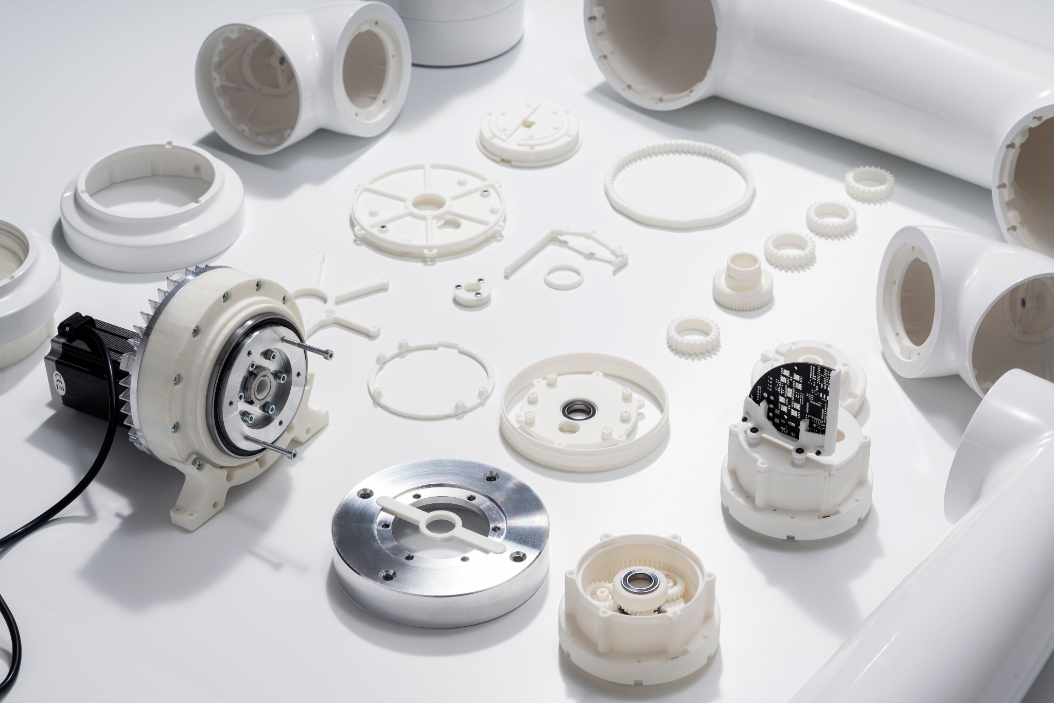 Różne białe elementy z tworzywa sztucznego i metalu, w tym rury, koła zębate i obudowy, rozłożone na białym tle.