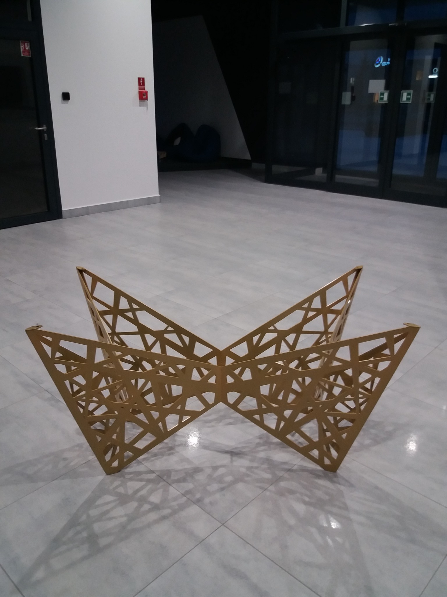 Geometryczna, ażurowa konstrukcja dekoracyjna z metalu w kształcie rozłożystego motyla, ustawiona na szarej podłodze w nowoczesnym wnętrzu.