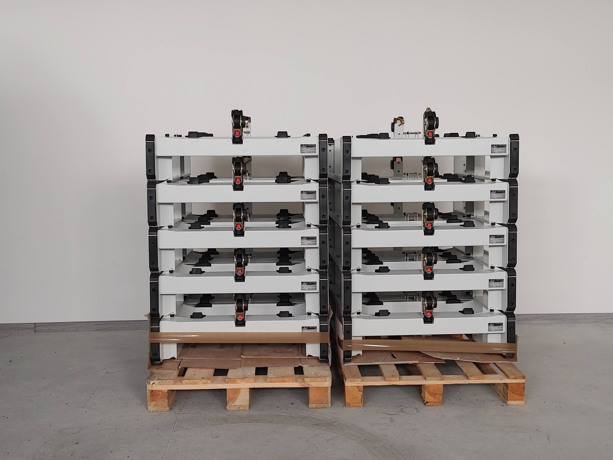 Dwa stosy białych, metalowych, modułowych konstrukcji na drewnianych paletach, gotowe do transportu, z widocznymi elementami mocującymi i etykietami producenta.