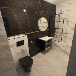 Nowoczesna łazienka z czarnymi i białymi płytkami imitującymi marmur, okrągłym lustrem w złotej ramie, czarną toaletą i umywalką na szafce z złotą armaturą.