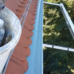 Malowany na jasnoniebiesko, metalowy system rynnowy zamontowany na dachu pokrytym czerwoną dachówką karpiówką, widok z góry z użyciem drabiny dekarskiej