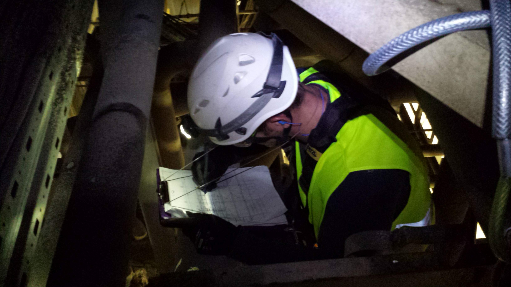 Pracownik w kasku i odblaskowej kamizelce, z lampką czołową, sprawdza dokumentację techniczną w trakcie inspekcji konstrukcji stalowej rurociągu w ciemnym otoczeniu przemysłowym.
