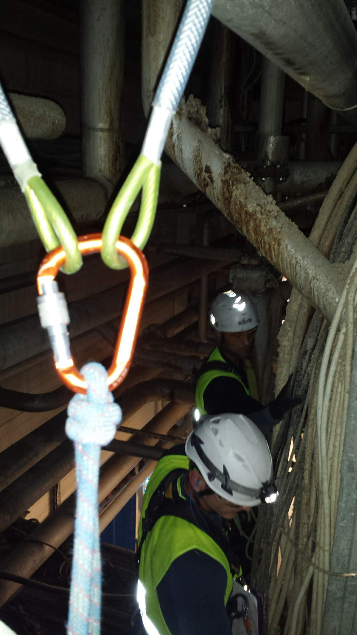 Dwóch pracowników w kaskach i kamizelkach odblaskowych, zabezpieczonych linami i karabińczykami, wykonuje inspekcję konstrukcji nośnej rurociągów w zakładzie produkcyjnym, widoczne zardzewiałe rury...