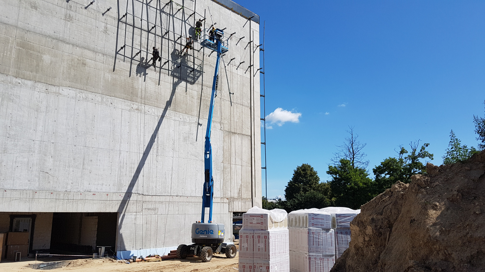 Montaż stalowej konstrukcji pod elewację wentylowaną na betonowej ścianie budynku z użyciem podnośnika Genie; widoczne materiały izolacyjne Rockwool i trzech pracowników na wysokości.