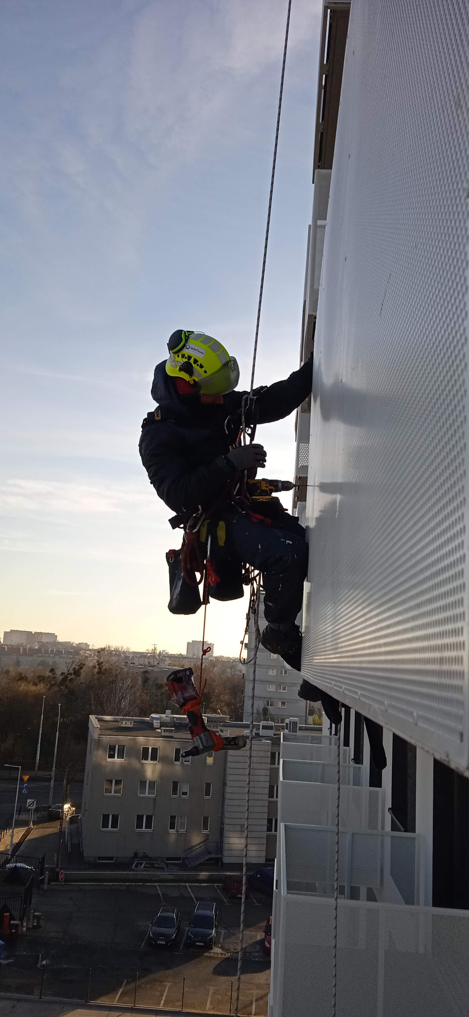 Pracownik na wysokościach montuje perforowaną blachę na balkonie, używając wkrętarki. W tle widoczny miejski krajobraz oświetlony wschodzącym słońcem.