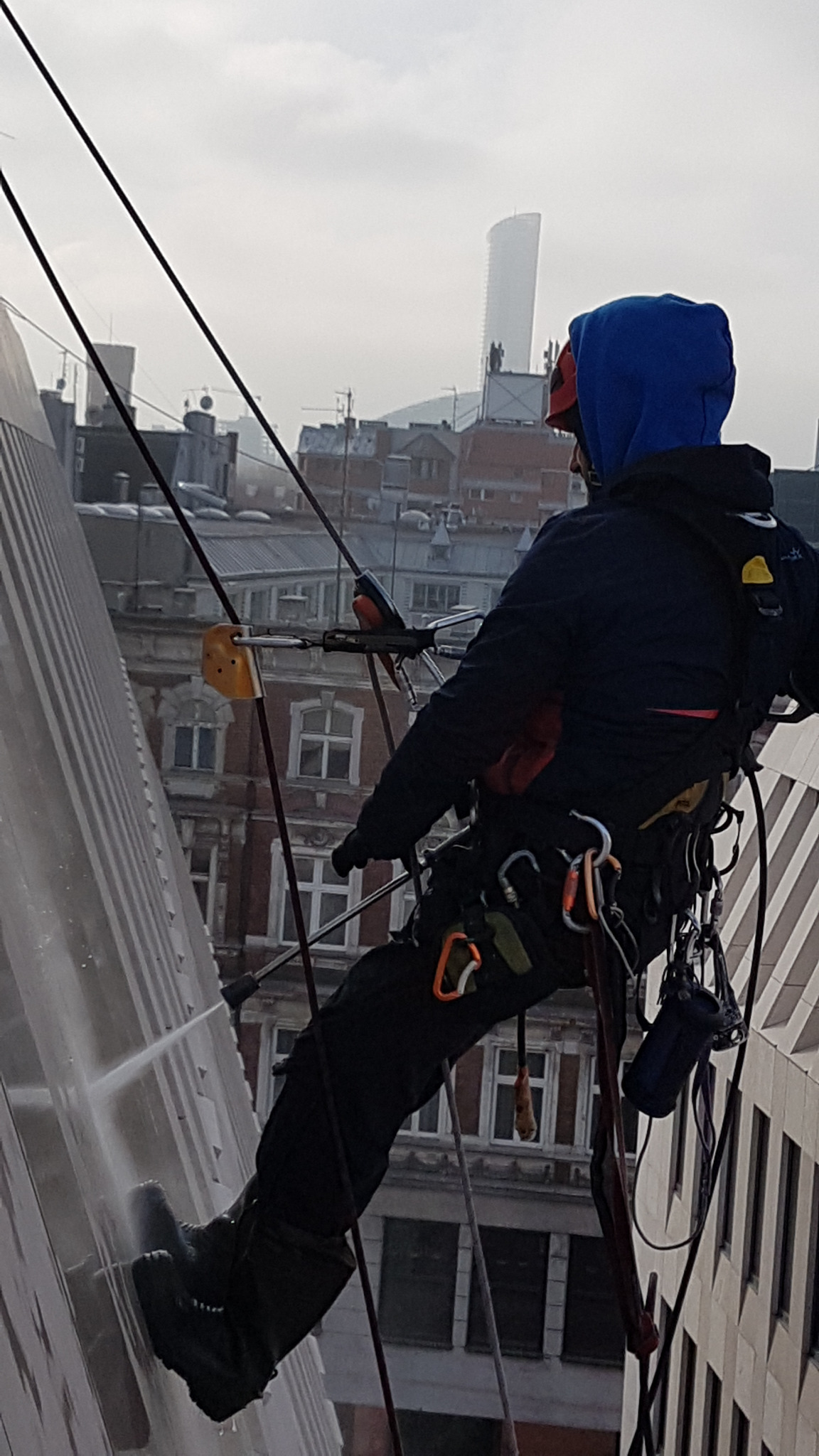 Pracownik w uprzęży alpinistycznej czyści elewację wysokiego budynku za pomocą myjki ciśnieniowej, w tle zabudowa miejska i wieżowiec.