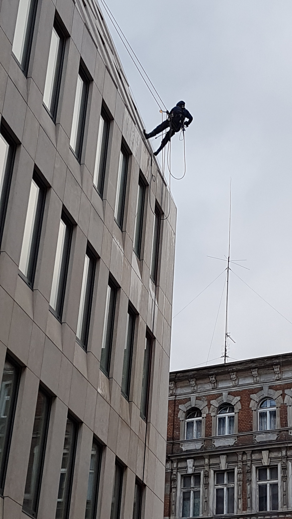 Pracownik na linach zabezpieczających, w kombinezonie, opuszcza się po elewacji nowoczesnego biurowca z licznymi oknami. W tle widoczny fragment starszej, ceglanej kamienicy z anteną.