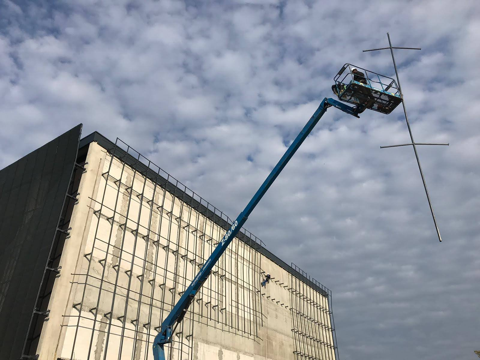 Pracownik na podnośniku koszowym montuje metalową konstrukcję na elewacji budynku w trakcie budowy, widoczne elementy mocujące i szkielet elewacji.