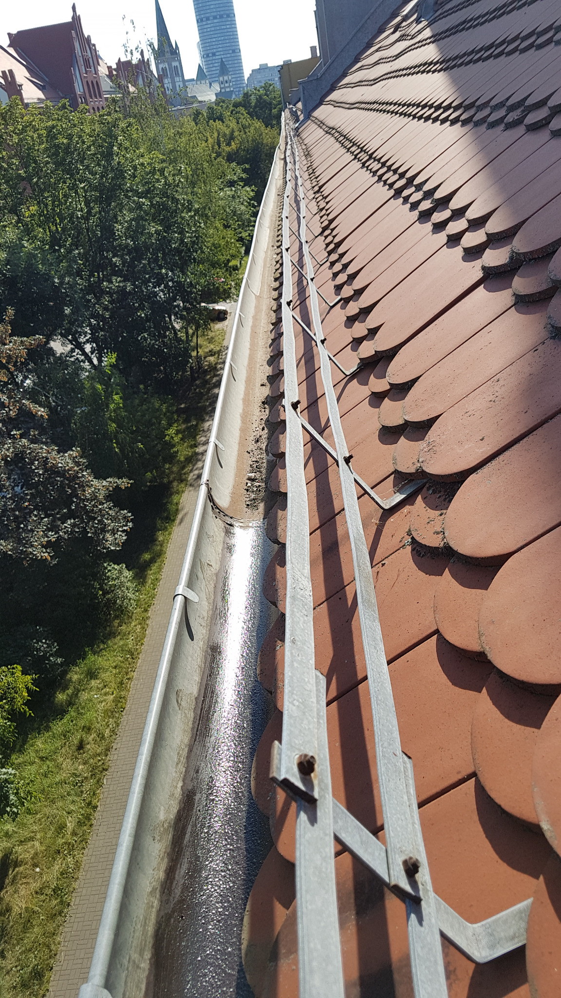 Częściowo zatkana rynna z wodą i zabrudzeniami, widok z góry na dach z czerwonej dachówki ceramicznej, w tle zabudowa miejska i zieleń.