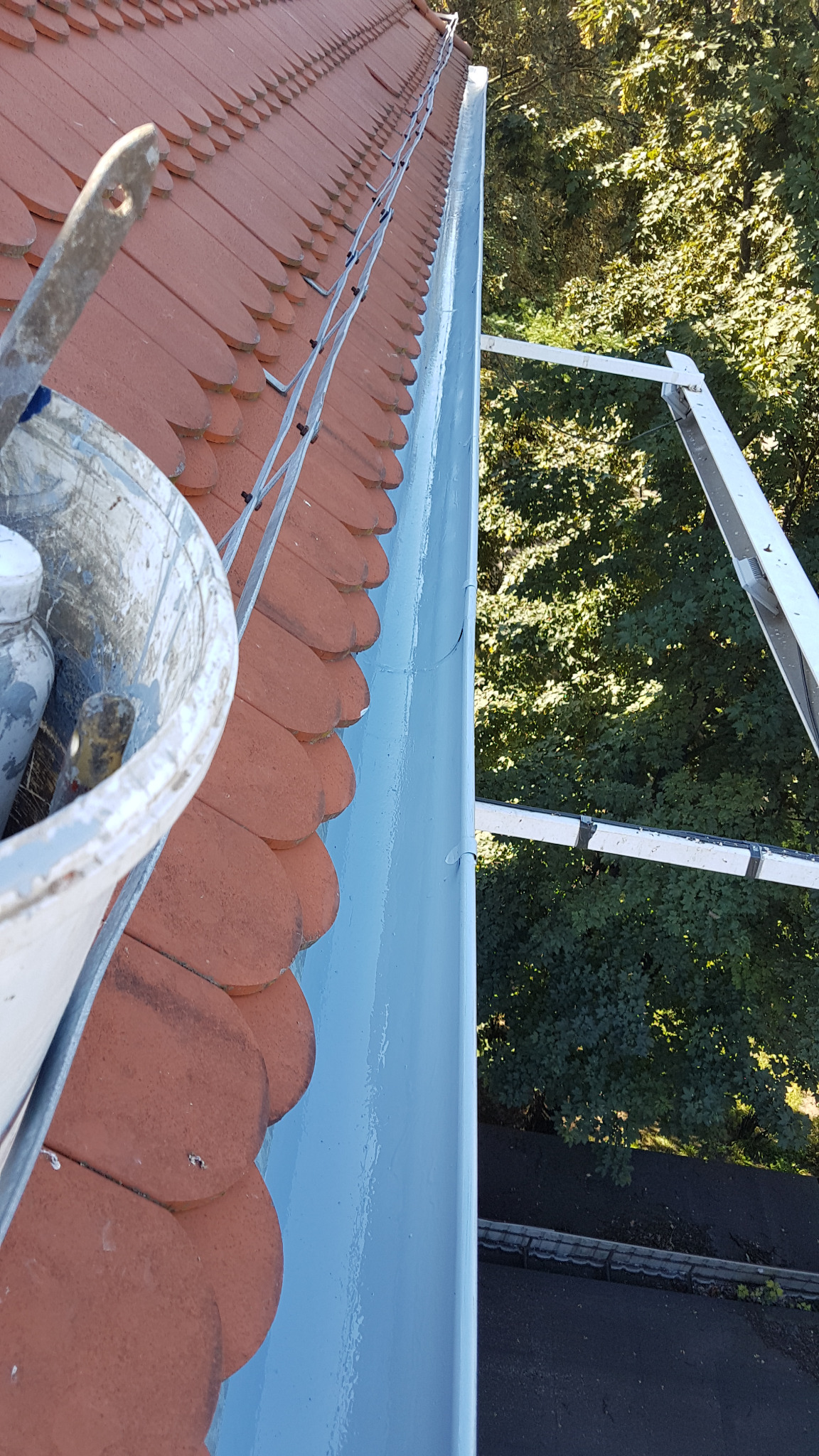 Malowany na jasnoniebiesko, metalowy system rynnowy zamontowany na dachu pokrytym czerwoną dachówką karpiówką, widok z góry z użyciem drabiny dekarskiej