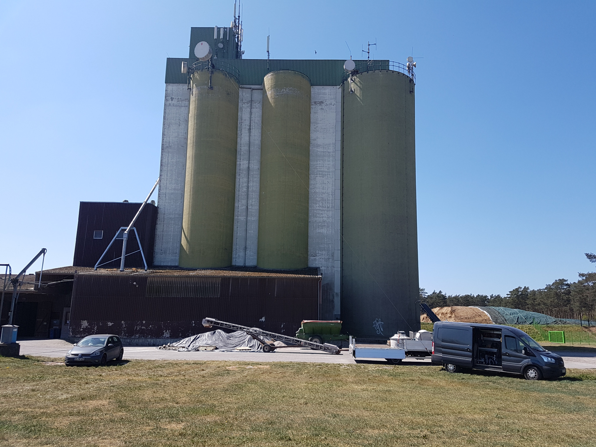 Widok silosów zbożowych z antenami na dachu, obok zaparkowane samochody i rozłożone urządzenia rolnicze