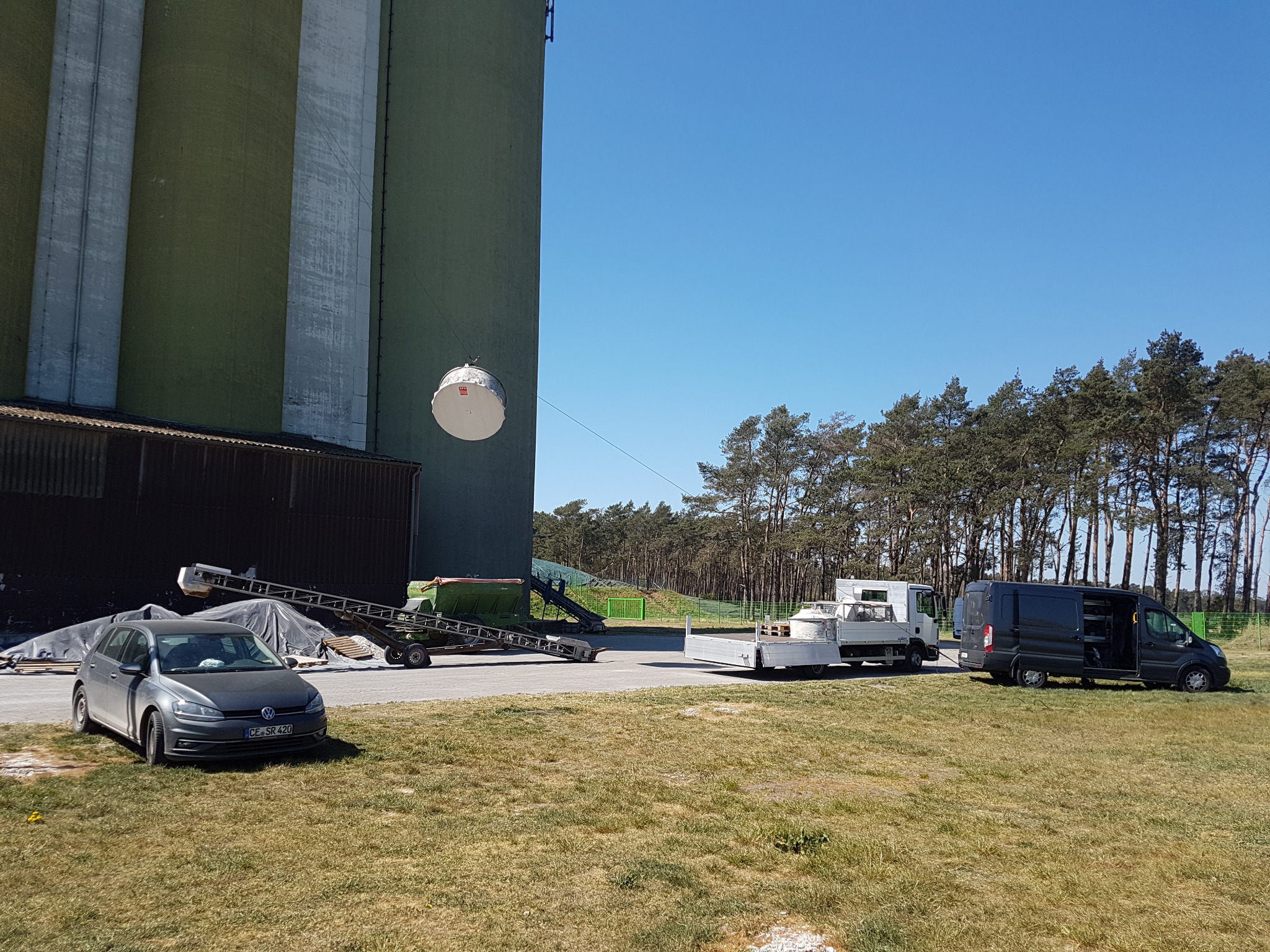Montaż okrągłej anteny na wysokim silosie, widoczny sprzęt i pojazdy na trawiastym terenie.