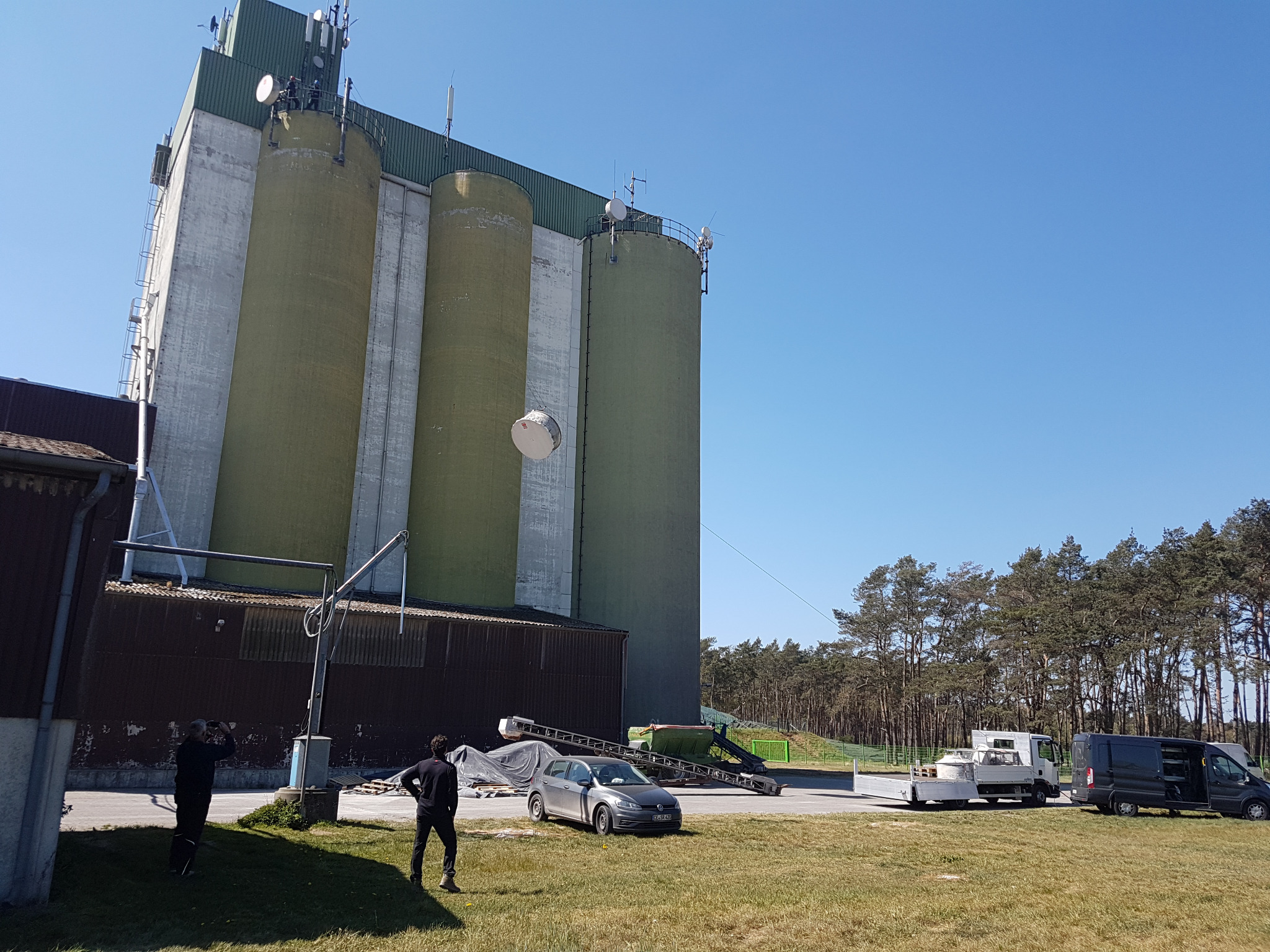 Wysoki silos zbożowy z zamontowanymi antenami, widoczny z poziomu gruntu, z pracownikami i samochodami na pierwszym planie. Na szczycie jednego z silosów widoczny pracownik w trakcie prac.