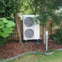 Biała, dwuczęściowa pompa ciepła na betonowych podstawach, stoi na korze drzewnej, otoczona zielenią i ceglanym murem. Widok z poziomu trawnika.